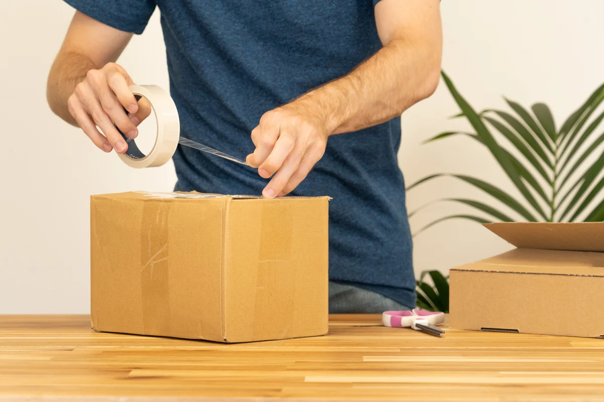 Shipper sealing a cardboard package with clear packing tape in preparation for weekend branch dispatch.